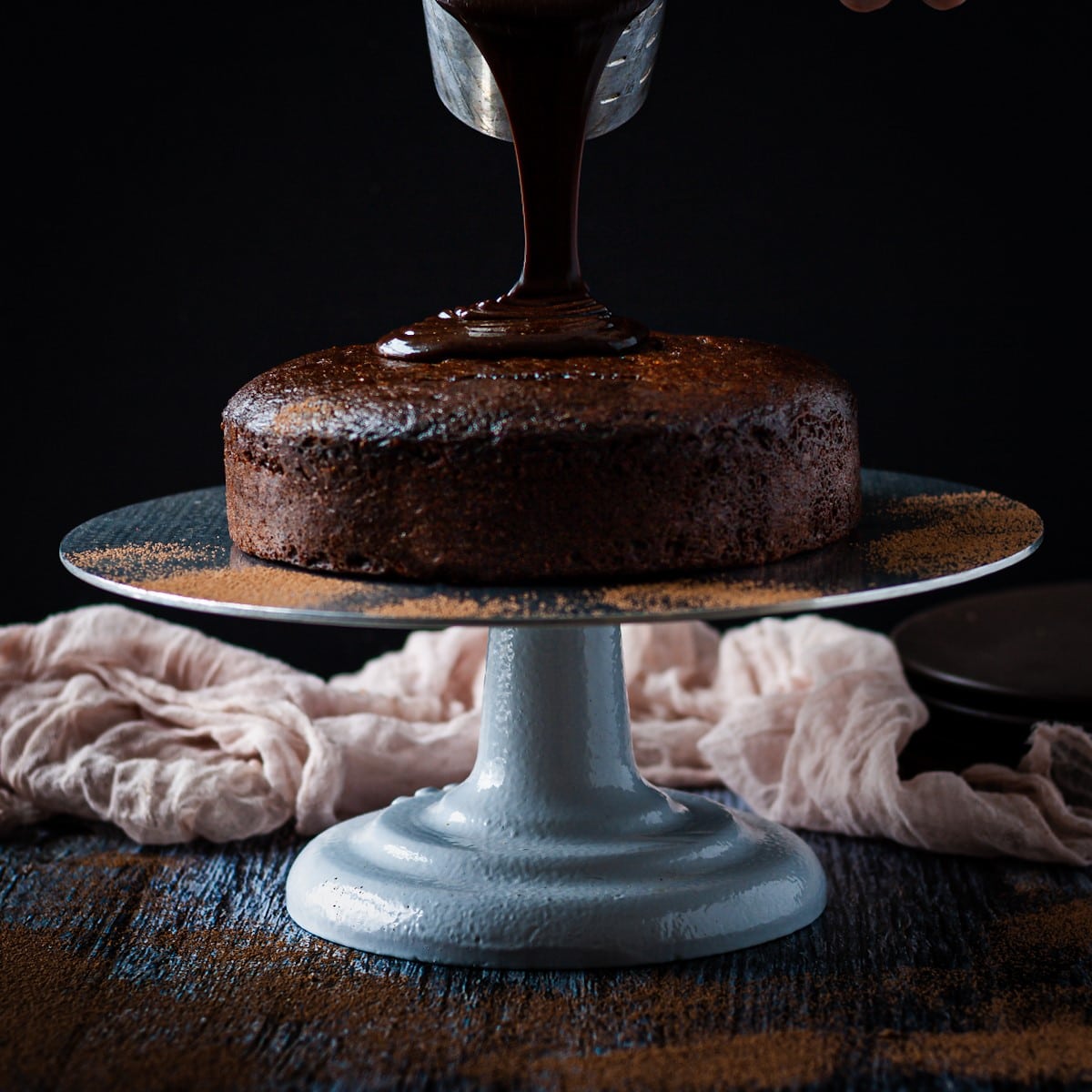 Ganache bring poured over a brownie cake on a cake stand.