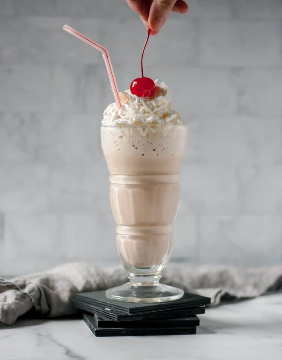 Nutella milkshake in a glass with whipped cream and a cherry being placed on top