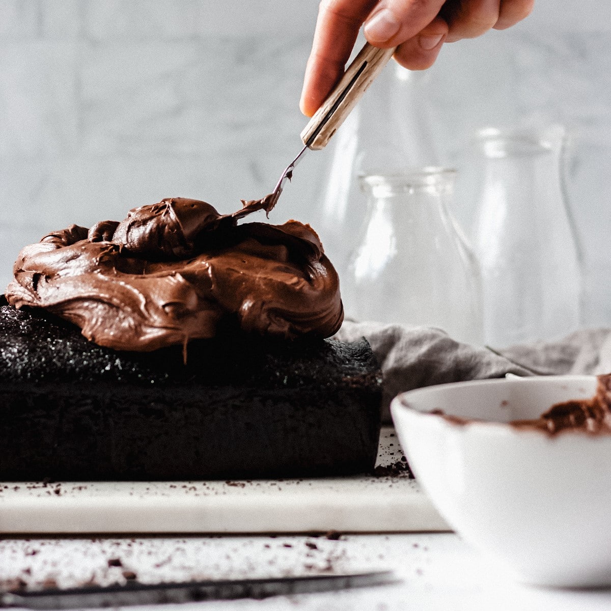 Chocolate loaf cake, sour cream fudge frosting being spread with a spatula and a small white bowl on the side.