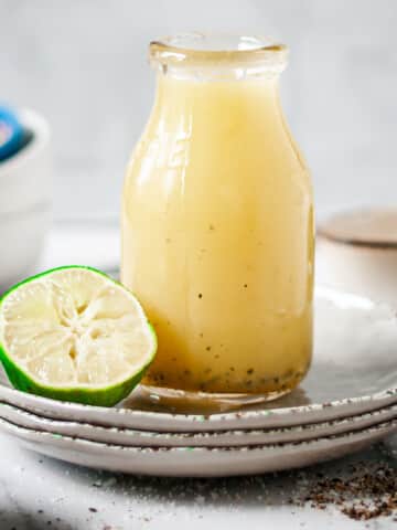 Glass jug of honey lime dressing on a stack of plates with a lime half.